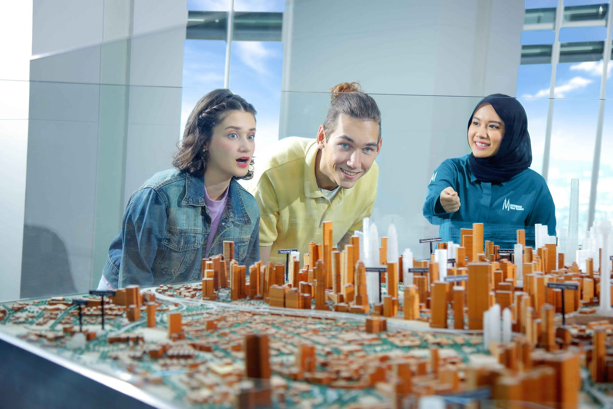 A PETRONAS Twin Towers guide pointing out landmarks on a large scale model of Kuala Lumpur to two visitors.