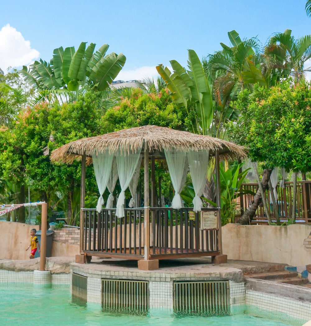 A thatched cabana with white curtains standing over a turquoise pool.
