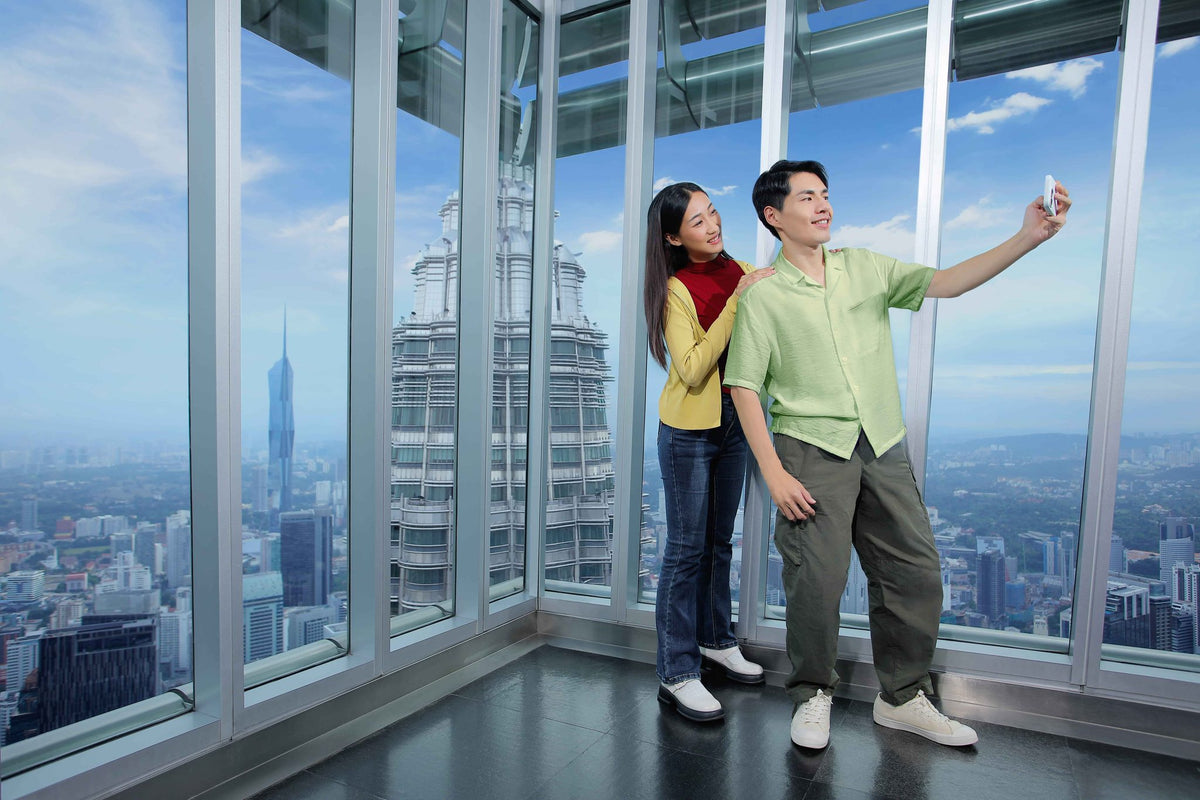 A couple taking a selfie at the Observation Deck window, with the second tower and the Kuala Lumpur skyline behind them.