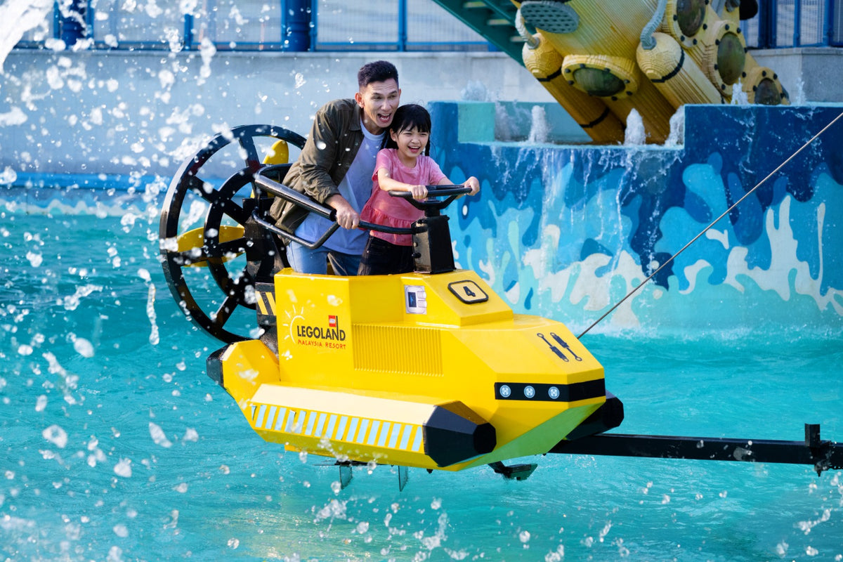 A man and a young girl steering a yellow LEGO submarine-styled boat through spraying water.