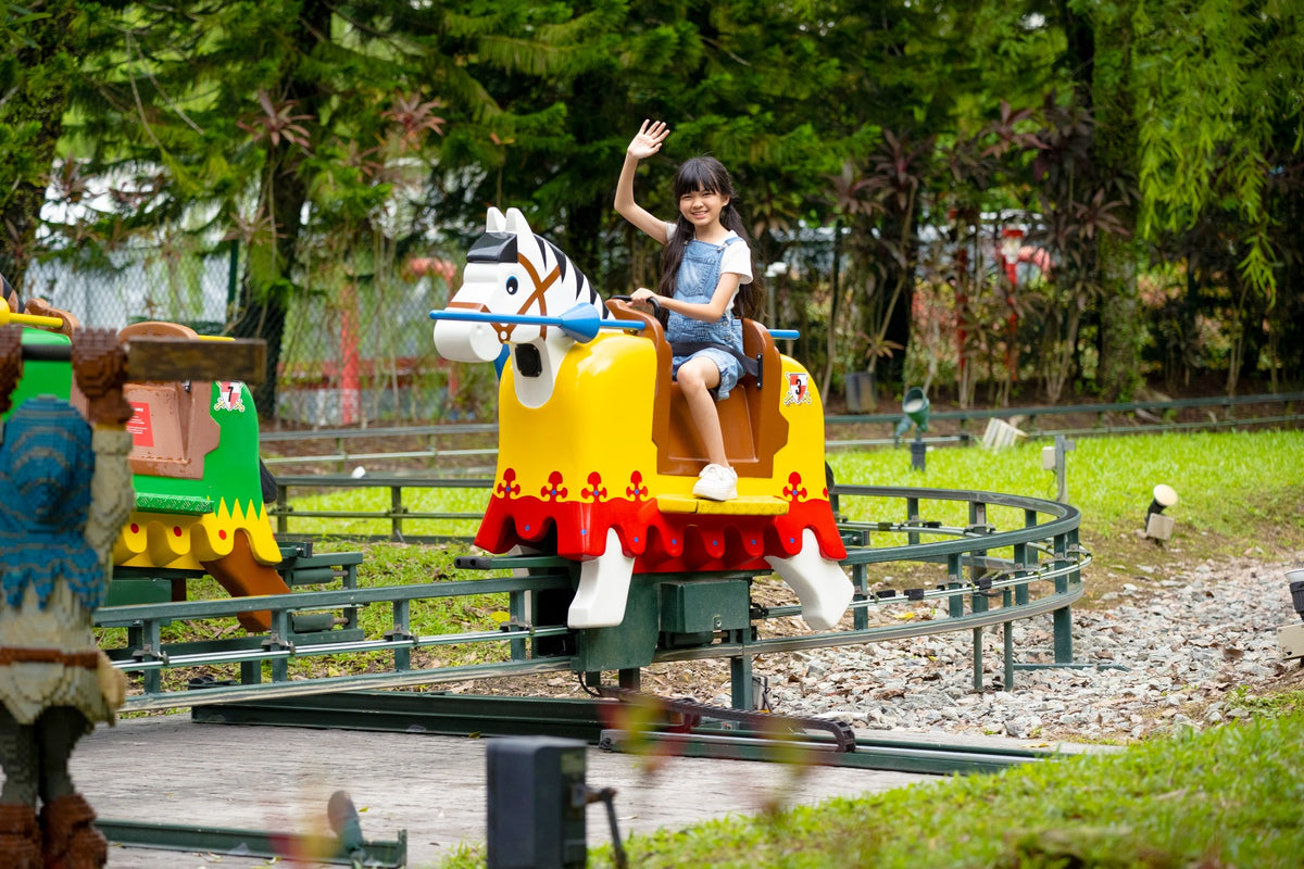 A girl waving from a yellow carousel horse on a small circular ride, with trees behind.