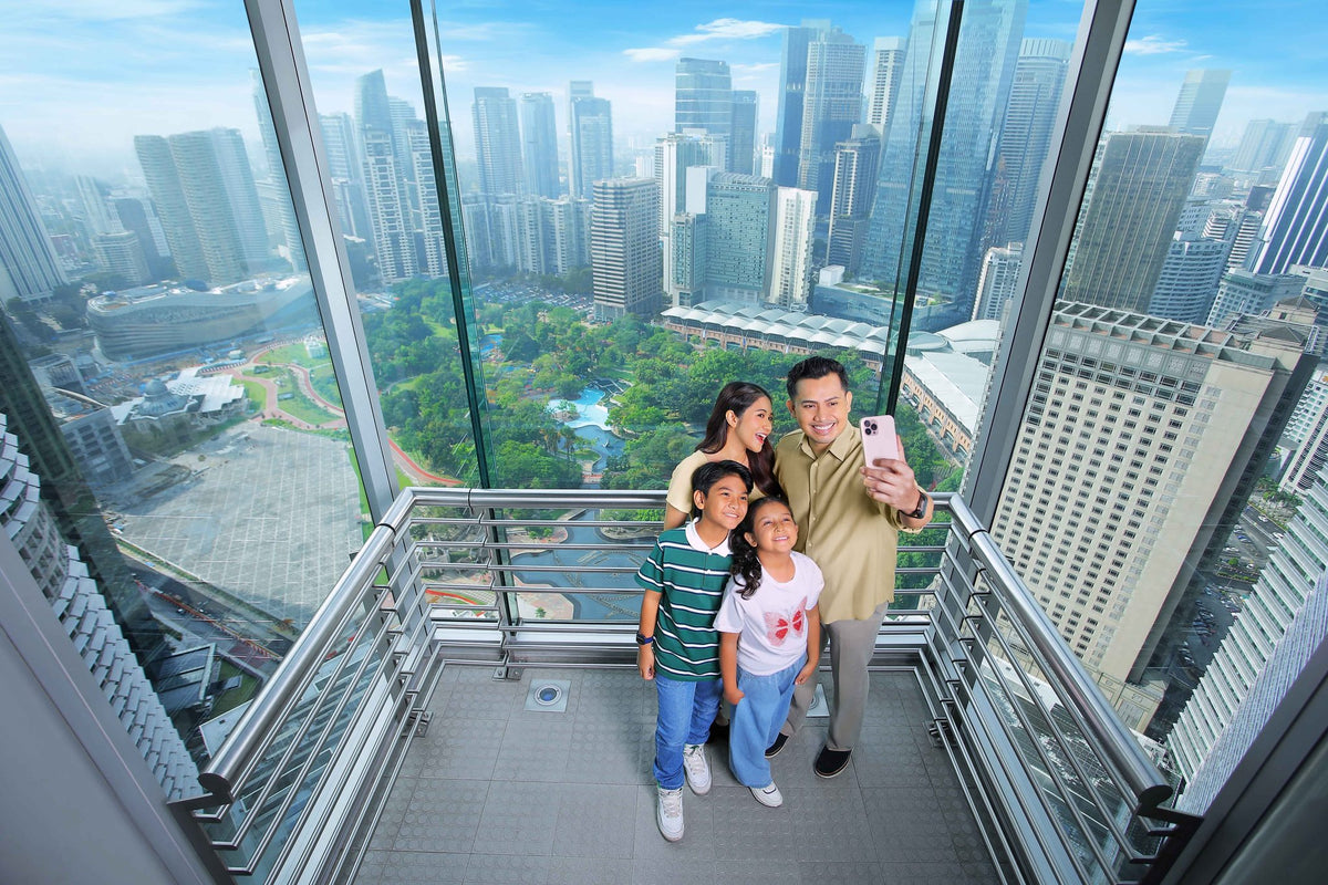 A family of four taking a selfie on the Skybridge with KLCC Park and the city spread out beneath them.