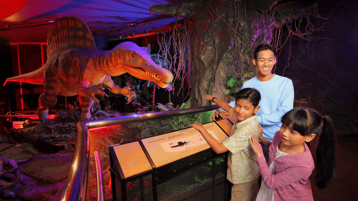 A family at an interactive display beside a life-size dinosaur model in a red-lit gallery.
