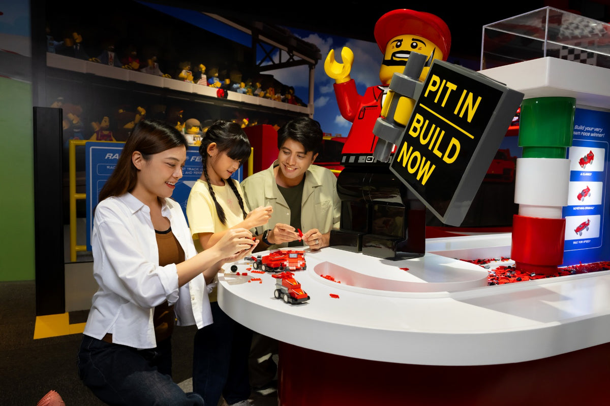 Three adults assembling LEGO Ferrari cars at a build station, beside a screen reading Pit In Build Now.
