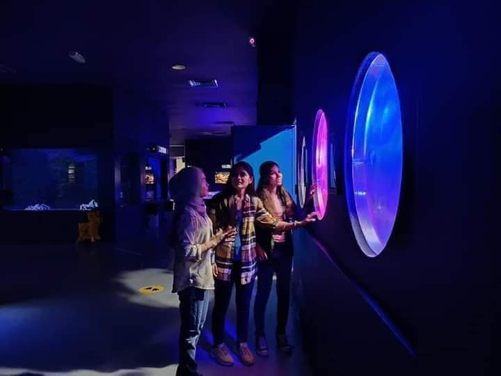Three visitors at a circular blue-lit porthole display in a darkened gallery.