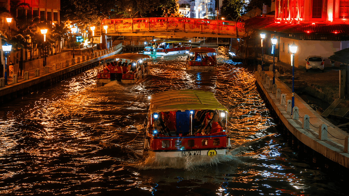 A cruise boat on the Melaka River at night, the banks lit orange and red.