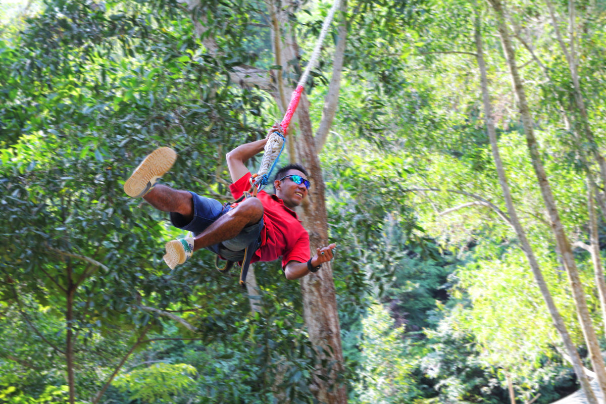 A man in a red shirt swinging through the forest canopy on a zipline harness.