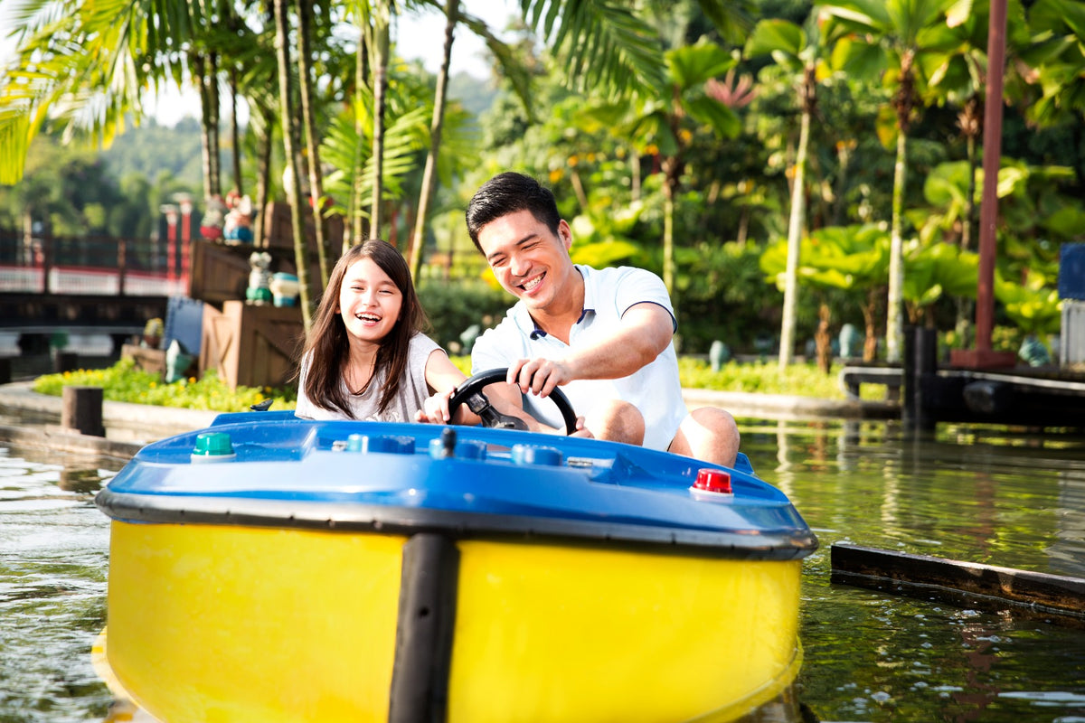 A man and a girl laughing at the wheel of a blue and yellow boat on a palm-lined lake.