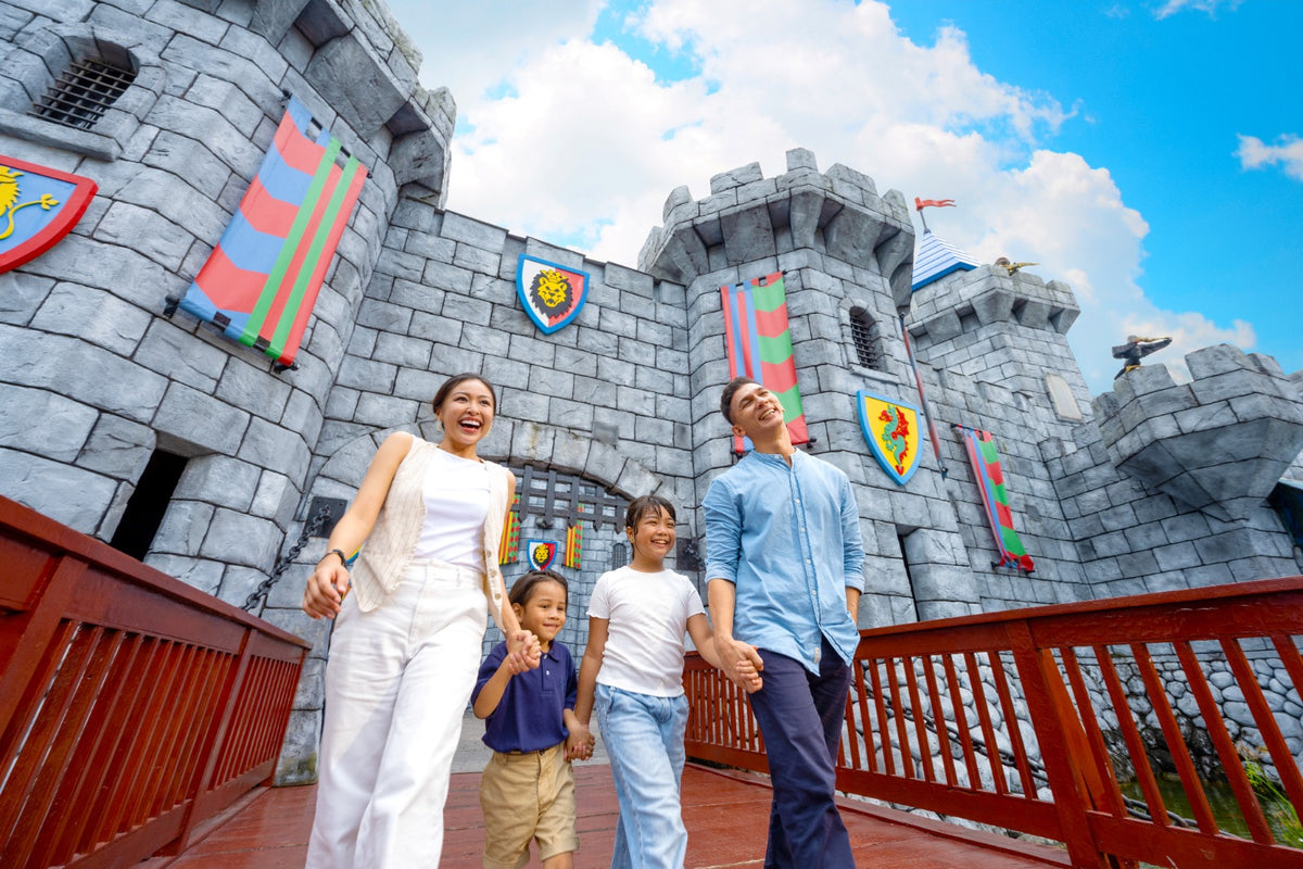 A family of four crossing a red wooden bridge beneath the grey LEGO castle walls, hung with heraldic banners.