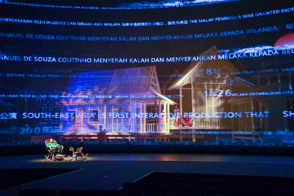 Projected historical text washing across the stage set of a wooden stilt house.