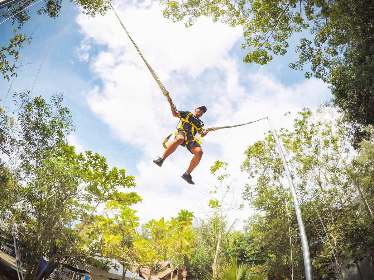 A visitor in a yellow harness suspended high against the sky on a bungee swing, seen from below.