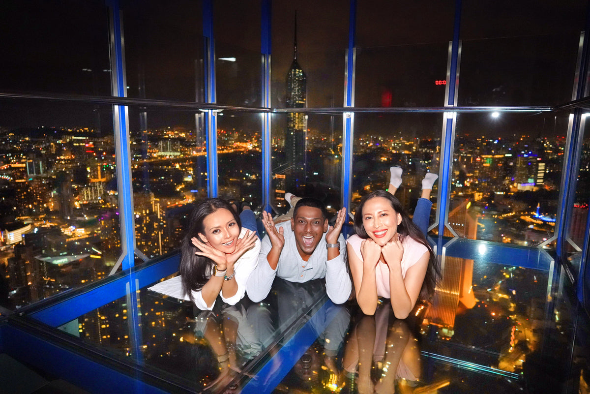 Three visitors lying on the glass floor of the Sky Box at night, city lights beneath them.