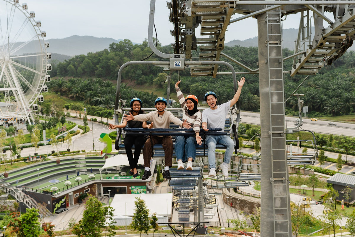 Four riders on the Skyride chairlift with their arms raised, a Ferris wheel and hills behind.