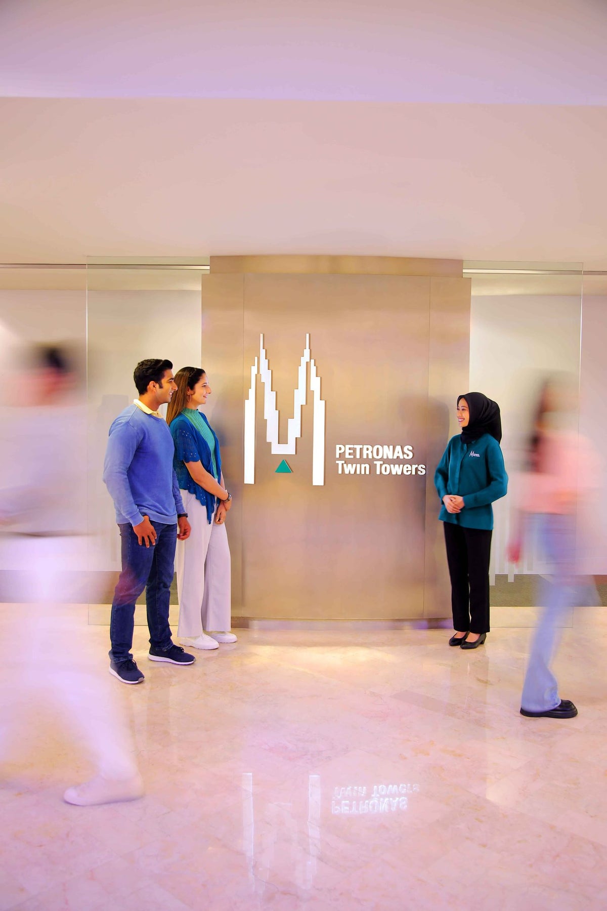 A PETRONAS Twin Towers host greeting two visitors beside the tower logo in the ground floor lobby.