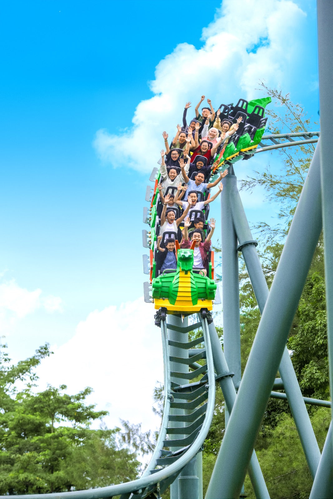 Riders with their arms in the air on the green dragon roller coaster, cresting a drop against a bright blue sky.