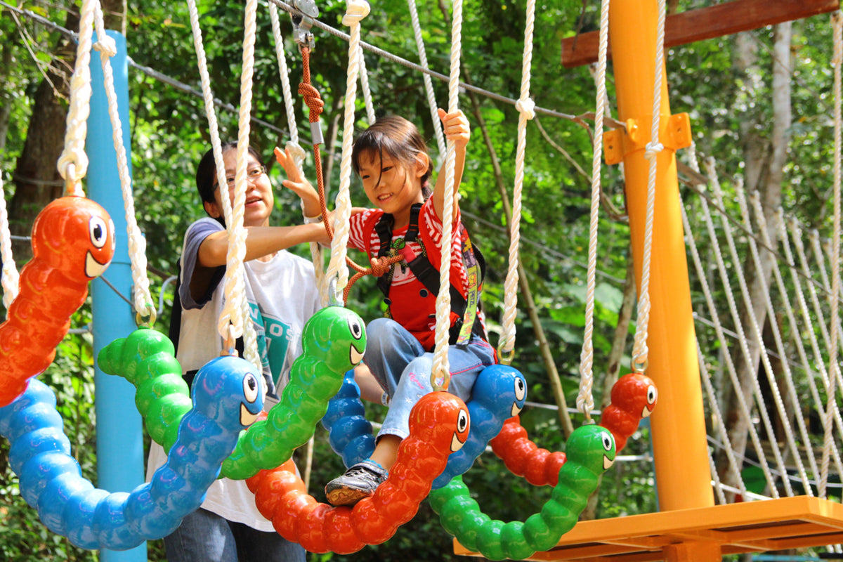 An adult and a child crossing a rope bridge strung with brightly coloured caterpillar floats.