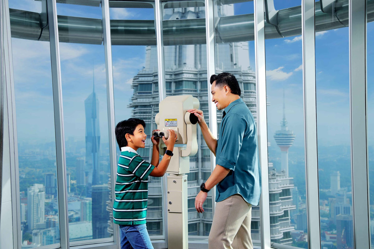 A boy using a viewing telescope on the Observation Deck while his father steadies it, with KL Tower in the distance.