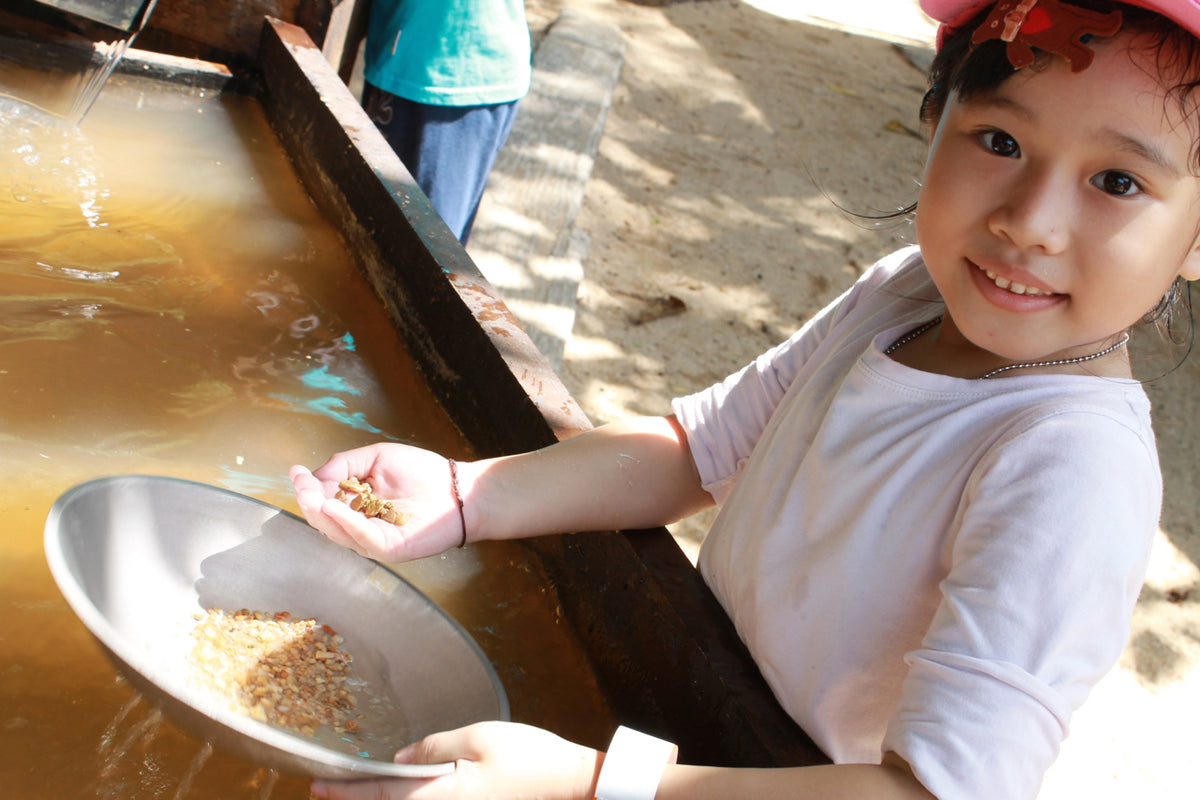 A girl panning for gold flakes in a metal dish at a water trough.