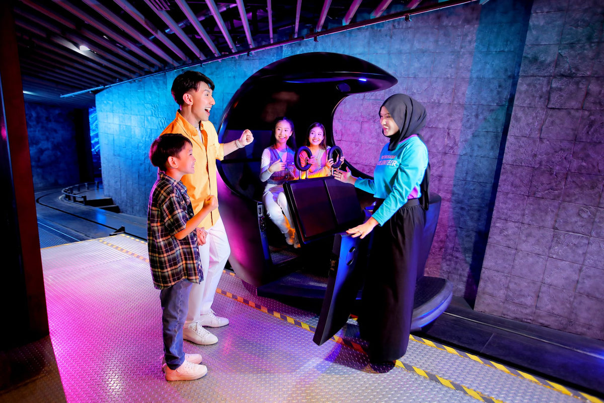 A Petrosains volunteer seating two girls in the Energy Capsule ride pod while a man and a boy wait their turn.