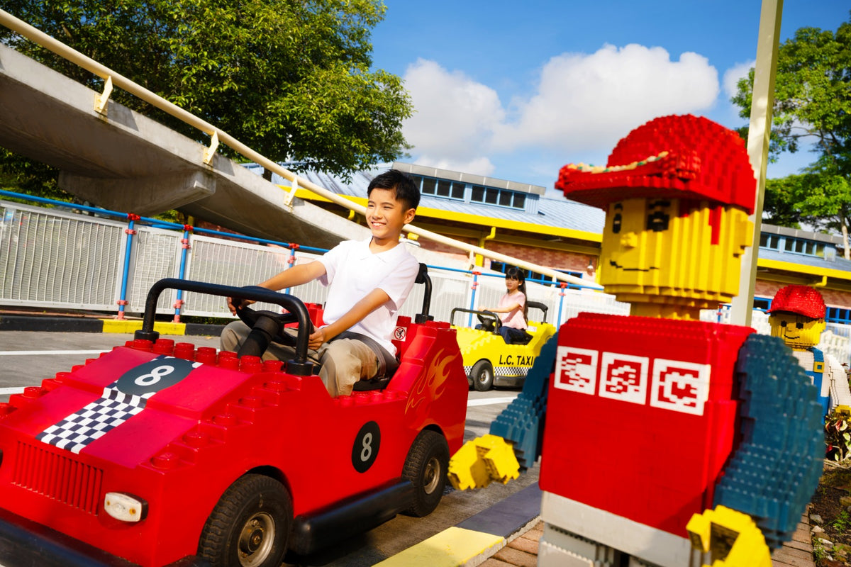 A boy grinning at the wheel of a red number 8 LEGO race car on the Driving School track, beside a large red LEGO minifigure model.