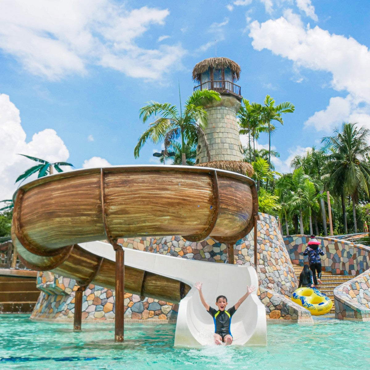 A boy with arms raised sliding into the pool beneath the lighthouse and wooden flume.