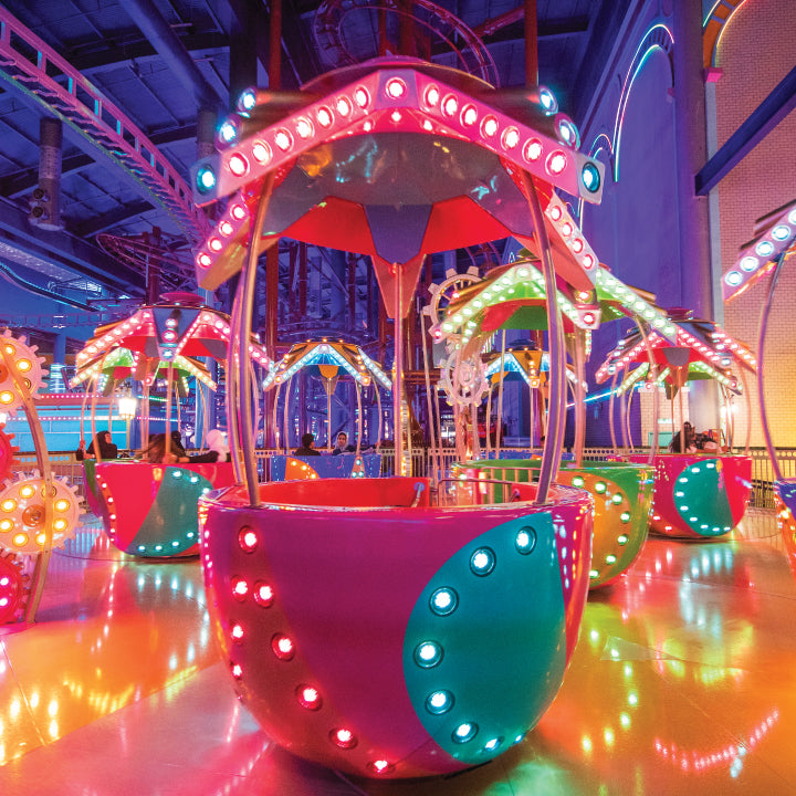 A brightly lit teacup ride spinning on the indoor park floor.