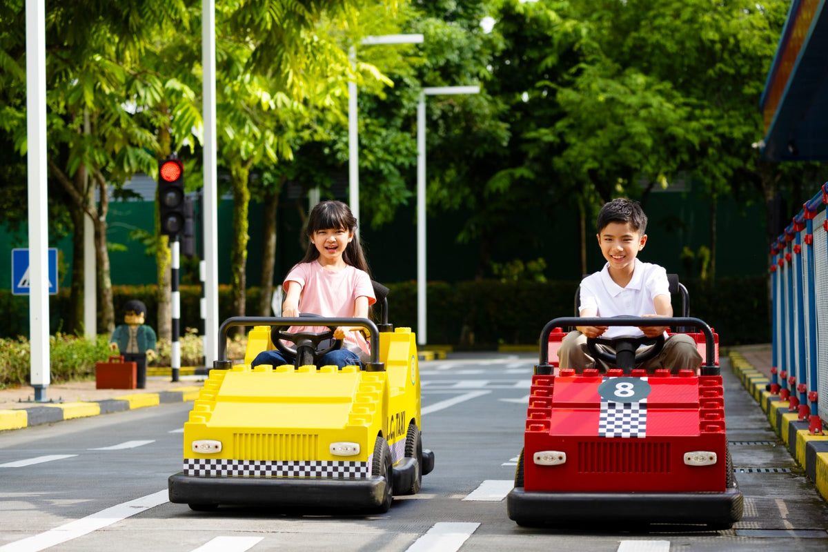 Two children driving a yellow and a red LEGO-bodied car on the Driving School circuit, with traffic lights and road markings.