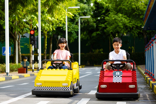 Two children driving a yellow and a red LEGO-bodied car on the Driving School circuit, with traffic lights and road markings.