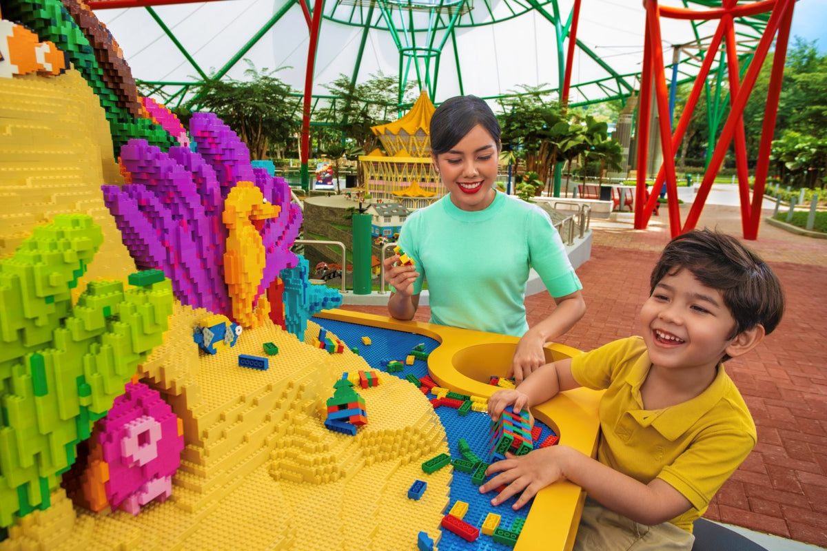 A woman and a boy building with LEGO bricks at a yellow play table, beside a large LEGO coral reef sculpture.