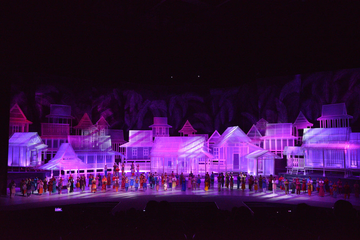 A stage filled with white model houses lit violet, with performers massed in front.