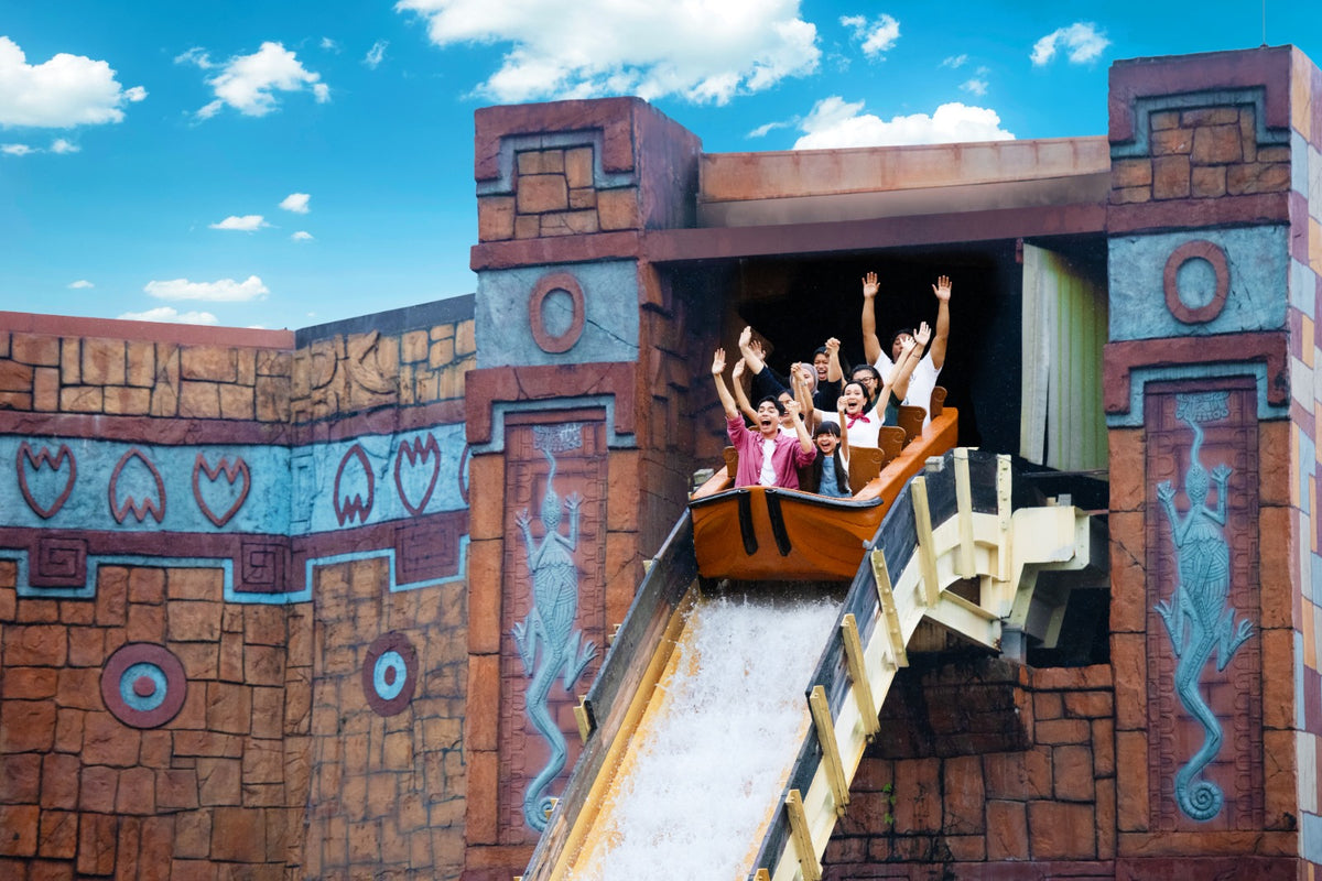A log flume boat plunging down the drop of a Maya-themed stone temple, riders with their arms raised.