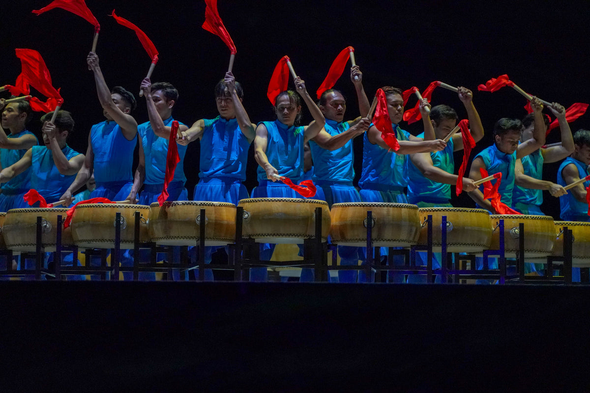 Drummers in blue costume raising red flags behind a row of large drums.
