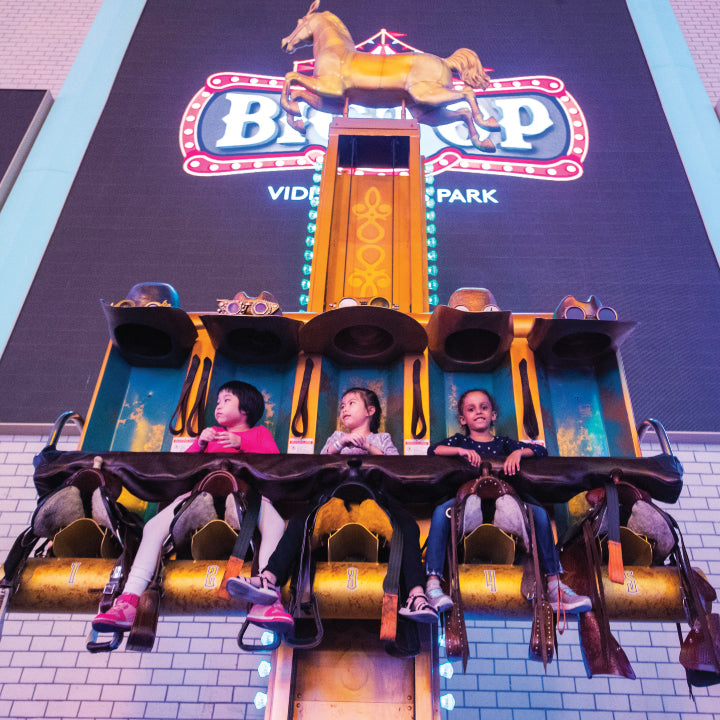 Three children riding a small indoor coaster past a painted backdrop.