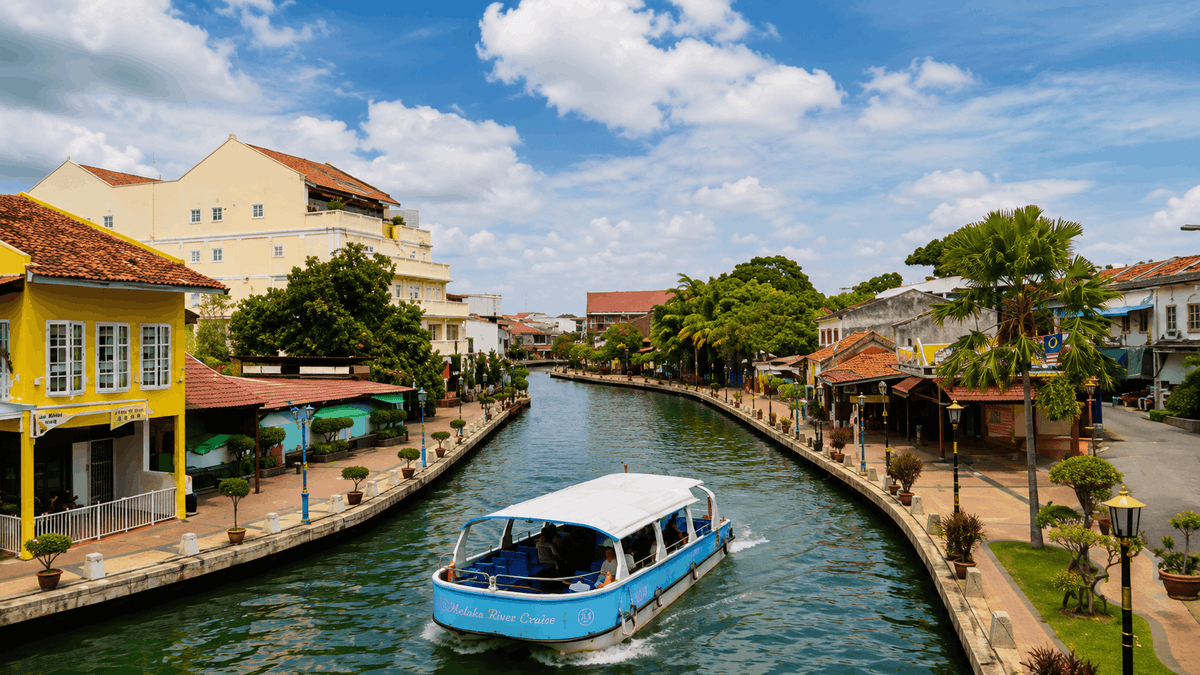 A white cruise boat passing colourful shophouses along the Melaka River in daylight.