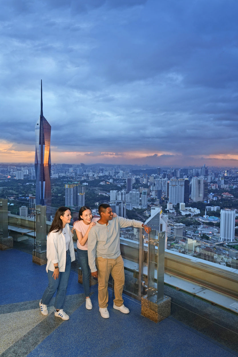 Three visitors at the Sky Deck rail at dusk, the city lights coming on below.