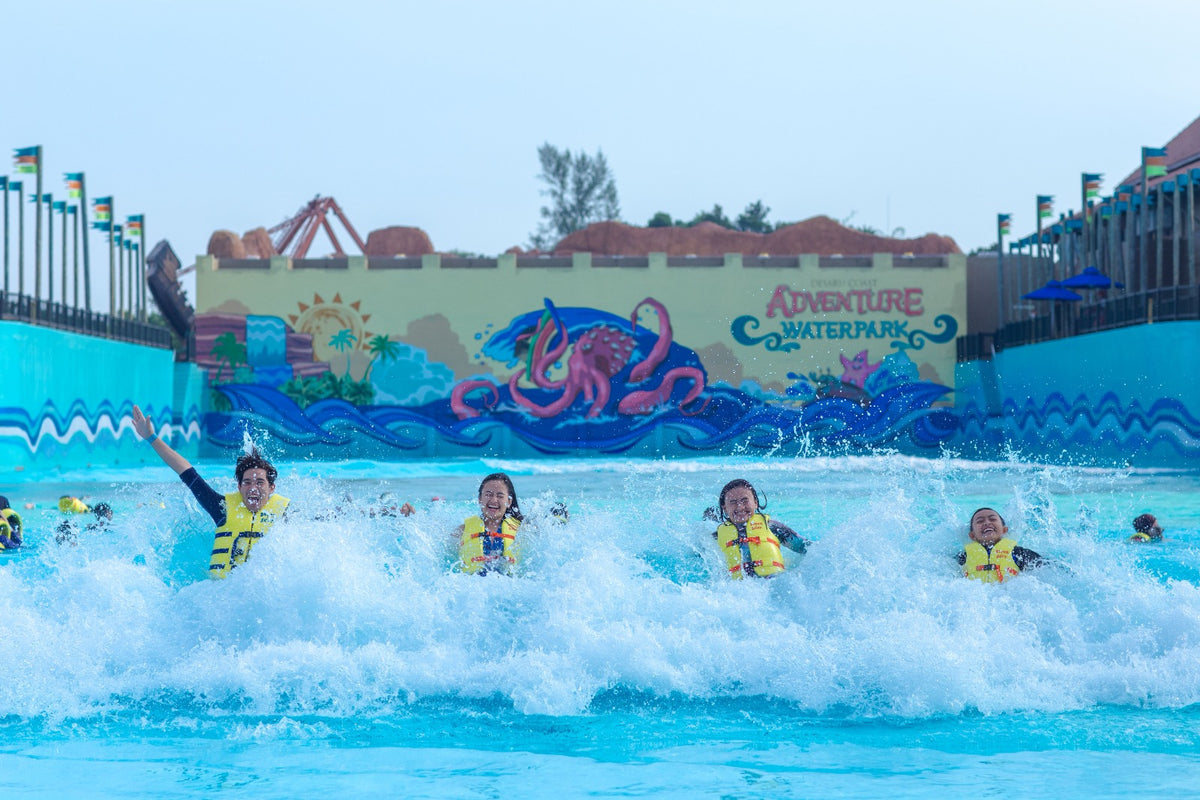 Children in life jackets riding the surf in the wave pool, with a painted octopus mural behind.