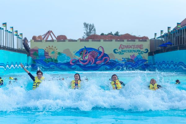 Children in life jackets riding the surf in the wave pool, with a painted octopus mural behind.