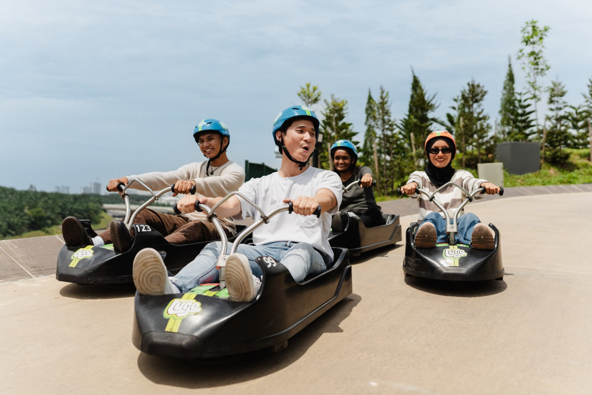 Four riders in helmets steering luge carts side by side down a paved track.