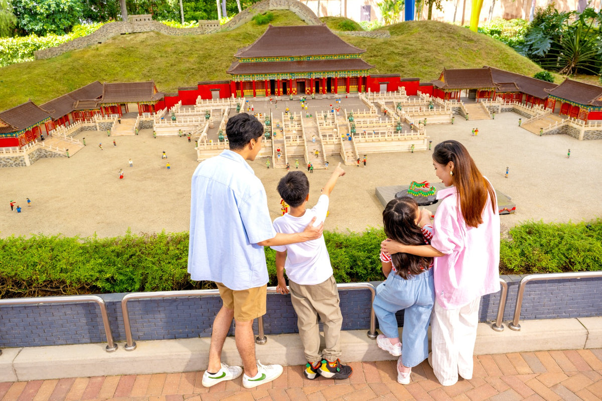 A family leaning on the rail above a MINILAND model of a red and white palace complex, a child pointing into it.