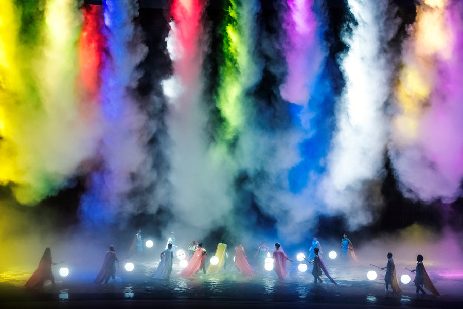 Performers on a flooded stage beneath columns of coloured smoke.