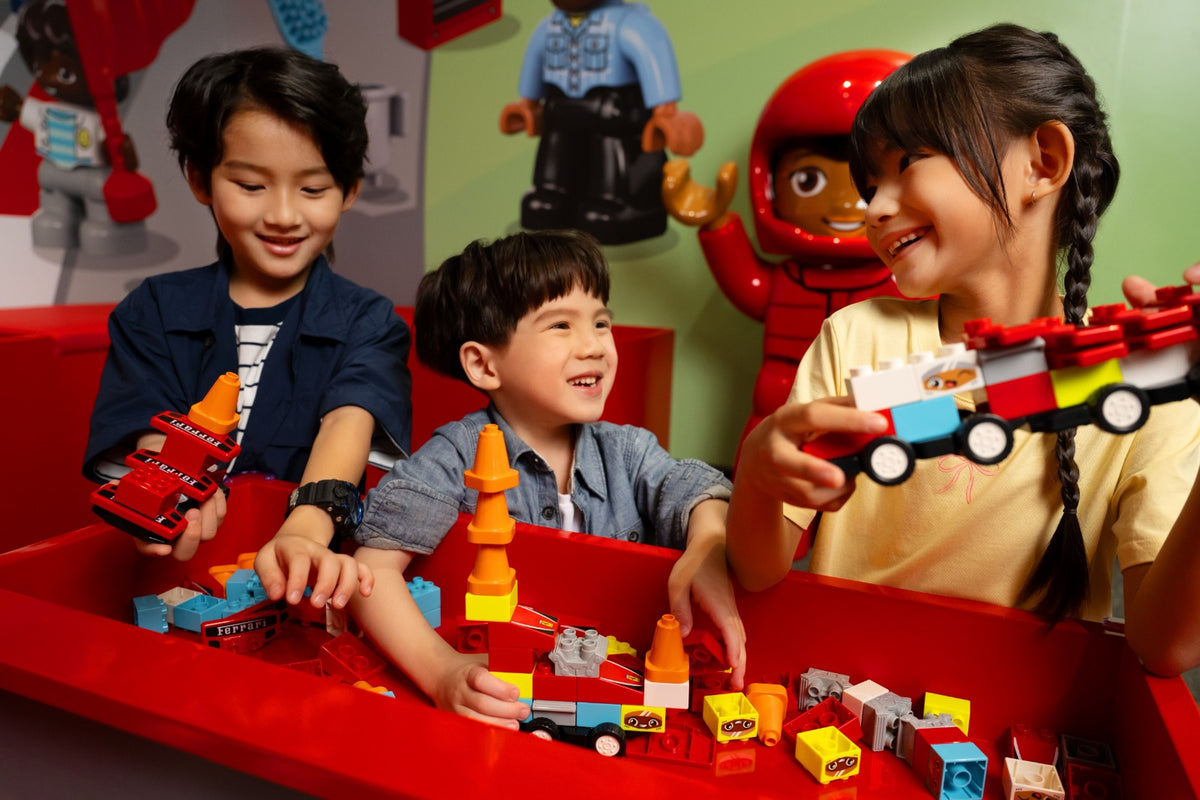 Three children building small LEGO cars at a red play table, with LEGO character models on the wall behind.
