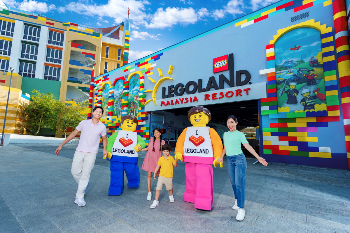 A family of four walking past the LEGOLAND Malaysia Resort entrance, flanked by two giant LEGO minifigure statues in I love LEGOLAND shirts.