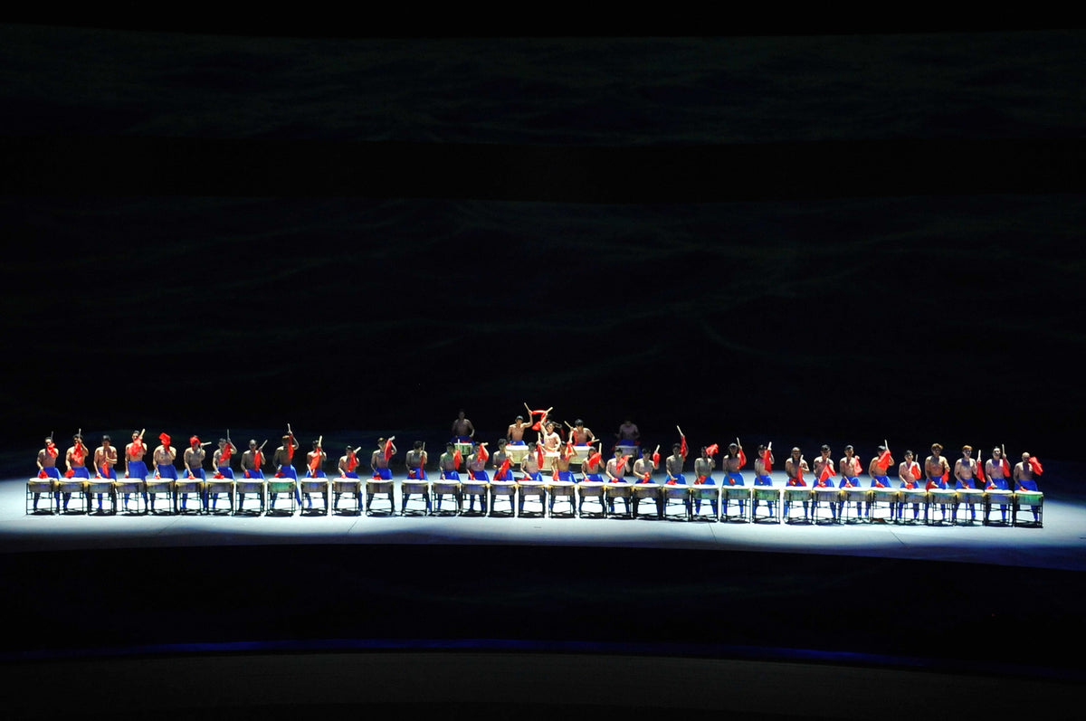 A long line of performers seated across a lit platform in the darkened auditorium.