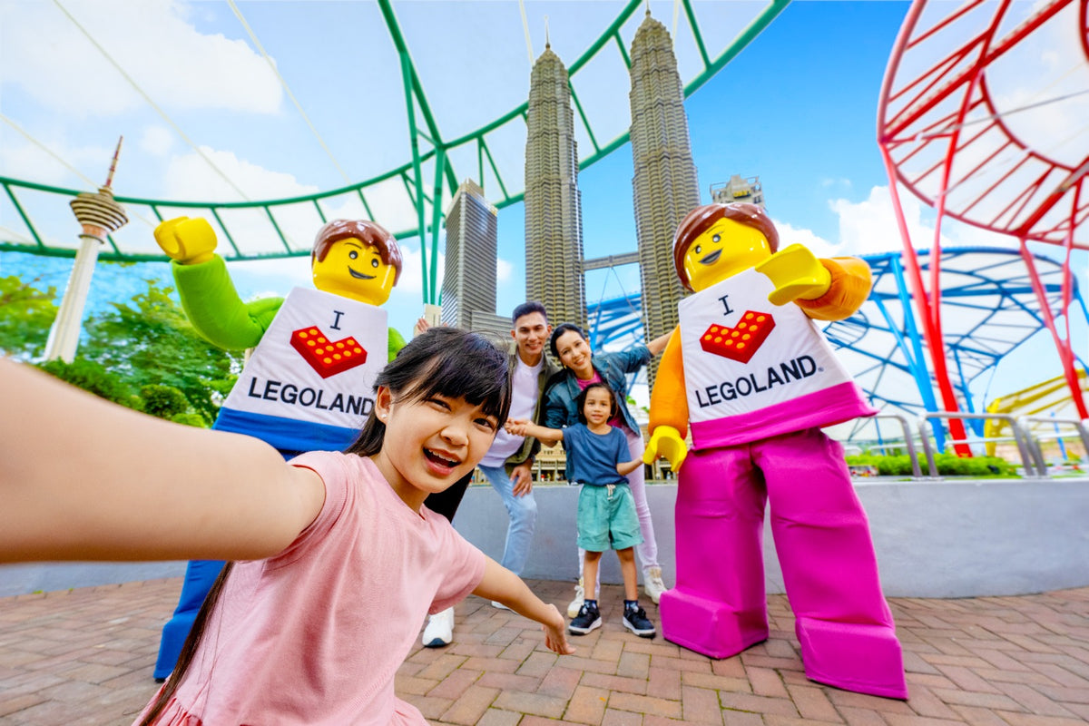 A family taking a selfie between two giant LEGO minifigures, in front of the MINILAND model of the Petronas Twin Towers.