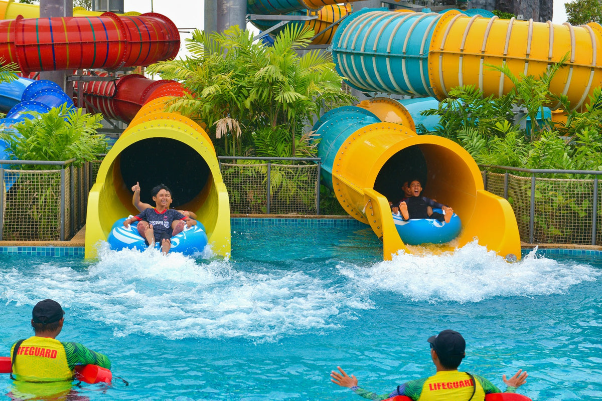 Two riders on tubes shooting out of yellow slide exits into the splash pool, lifeguards watching.
