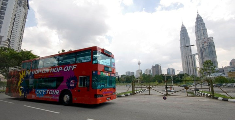 A red open-top tour bus on the road with the Petronas Twin Towers and KL Tower behind.