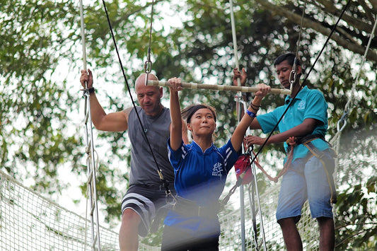 Two visitors balancing along a rope-and-pole obstacle course while an instructor steadies the line.
