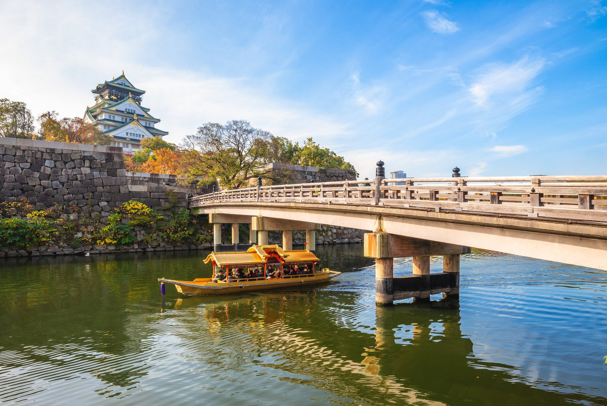 A traditional gold-roofed boat passing under a stone bridge on the Osaka Castle moat, with the keep behind.