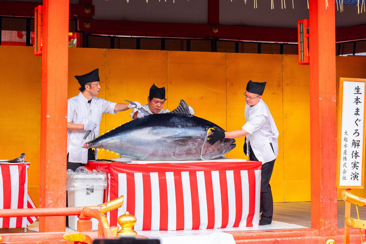 A tuna-cutting demonstration on a red and white draped stage, watched from beneath vermilion shrine pillars.