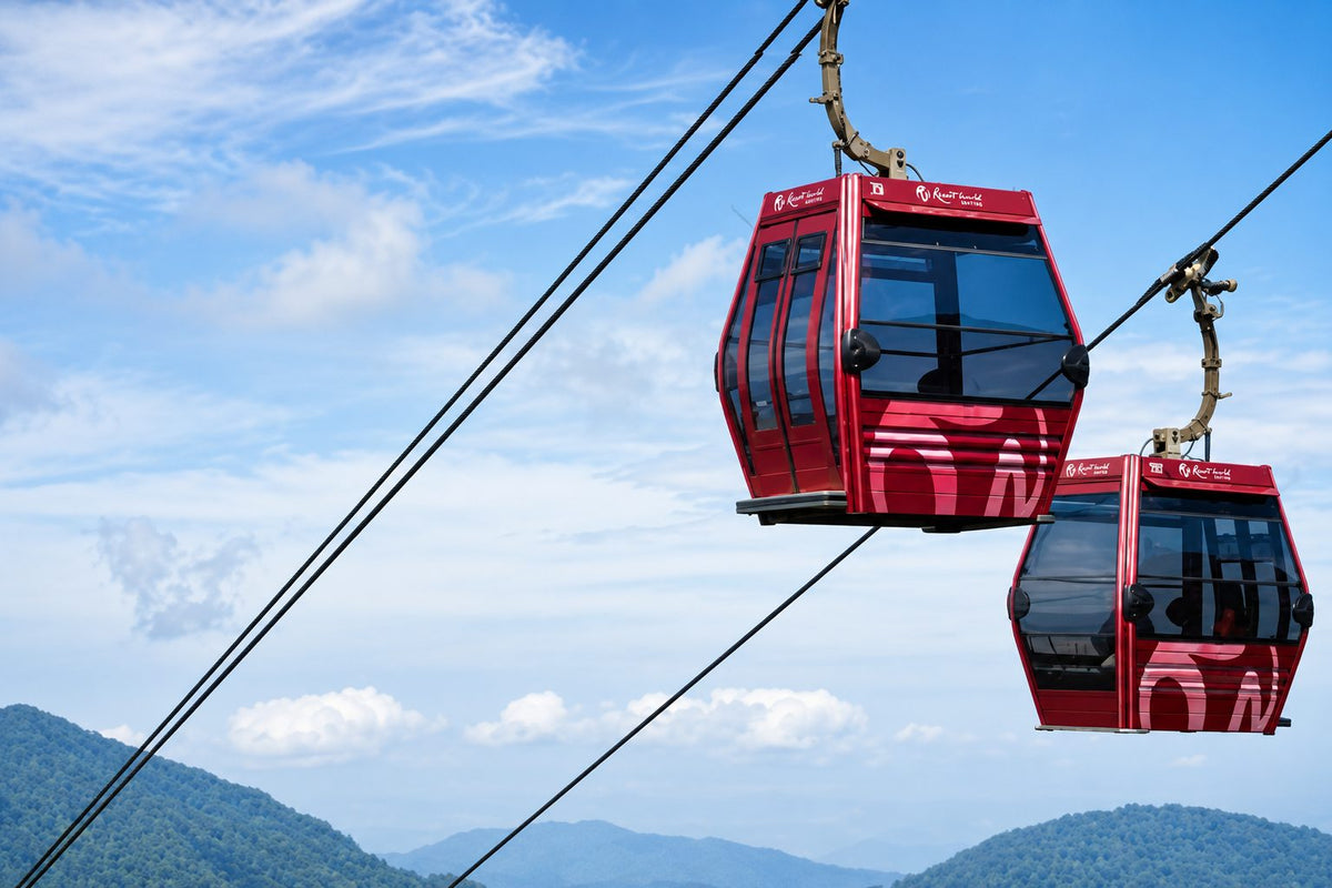 Two red Resorts World Genting cable car gondolas on the line against a blue sky, with forested hills below.
